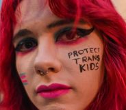 Young white person with red hair and 'protect trans kids' written on their cheek