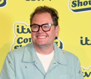 Alan Carr in a mint green shirt and white t-shirt smiling and standing in front of a yellow background.