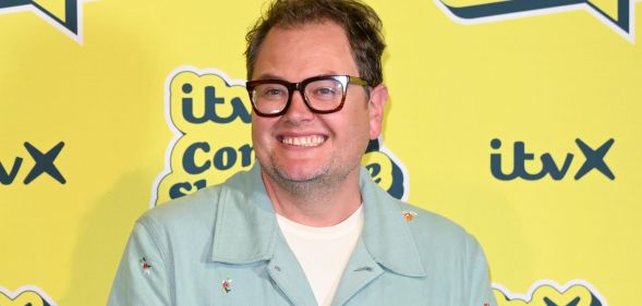 Alan Carr in a mint green shirt and white t-shirt smiling and standing in front of a yellow background.