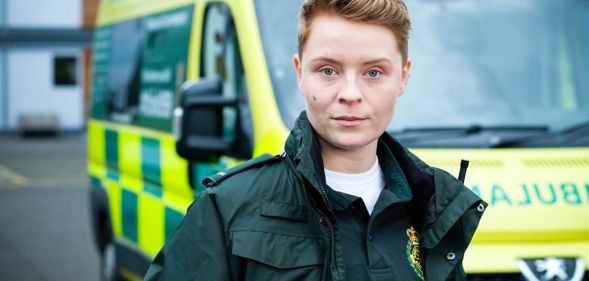 Actor Arin Smethurst as Casualty character Sah. Sah wears a green paramedic's outfit and stands against a UK ambulance.