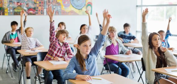 Children sit in a classroom holding their hands up