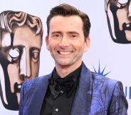 David Tennant wears a blue suit and black shirt while smiling at the camera.