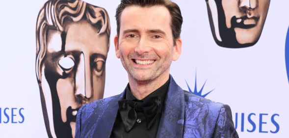 David Tennant wears a blue suit and black shirt while smiling at the camera.
