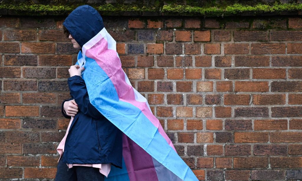A hooded individual with a trans flag wrapped around them walks in the rain.