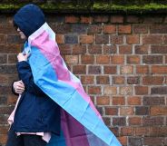 A hooded individual with a trans flag wrapped around them walks in the rain.