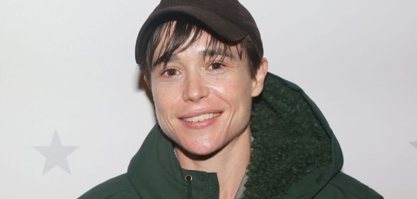 Elliot Page wears a thick green coat and brown cap while smiling at the camera.