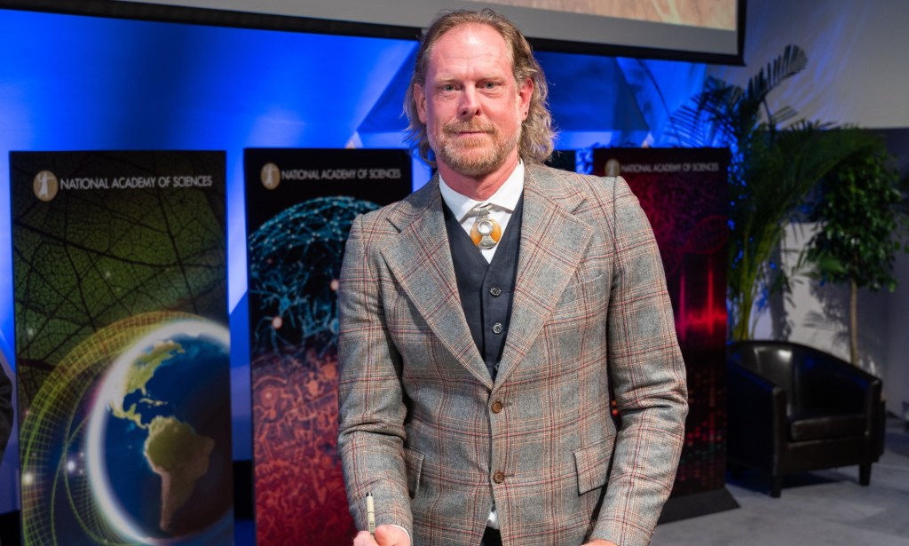 This is an image of Professor Giles Oldroyd. He is wearing a grey tweed suit and is standing in front of signs. He has a short beard and long brown-ish red hair.