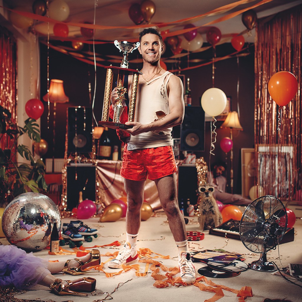 Jake Shears' Last Man Dancing album cover featuring the singer in a white vest and red shorts holding a trophy standing in a messy room after a party.