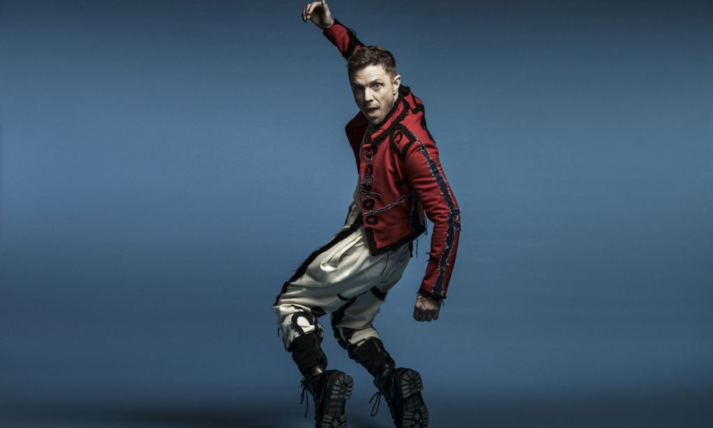 Jake Shears poses for a promo photo for his album Last Man Dancing. He is wearing silver trousers and a red jacket and standing against a blue background.