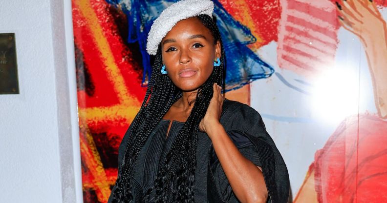 US singer and actor Janelle Monáe wears a black top, white beret and blue earring while standing against a red, orange, white and blue background.
