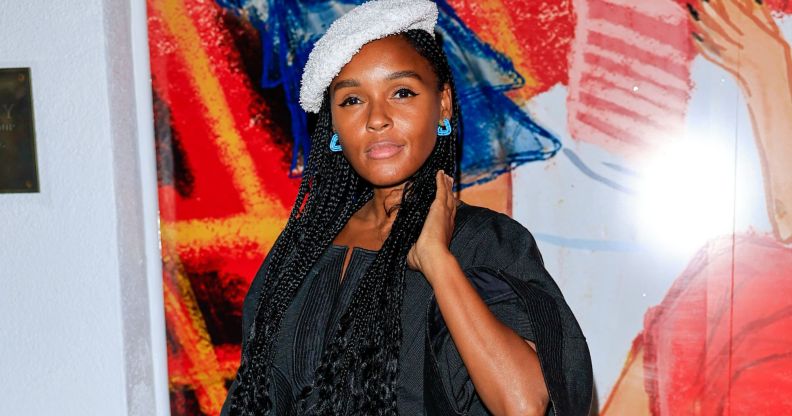 US singer and actor Janelle Monáe wears a black top, white beret and blue earring while standing against a red, orange, white and blue background.