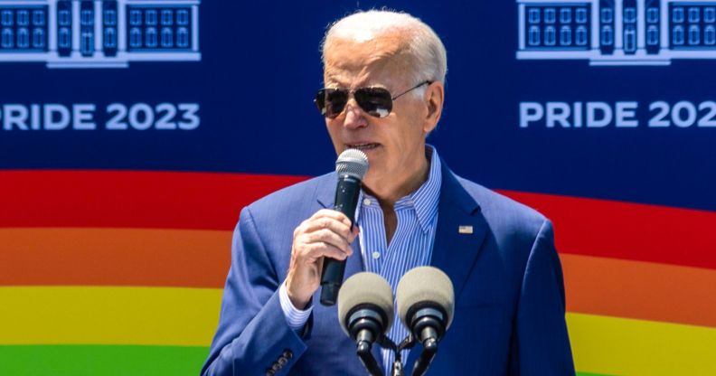 President Joe Biden speaking into a microphone, with a picture of a rainbow flag behind him.