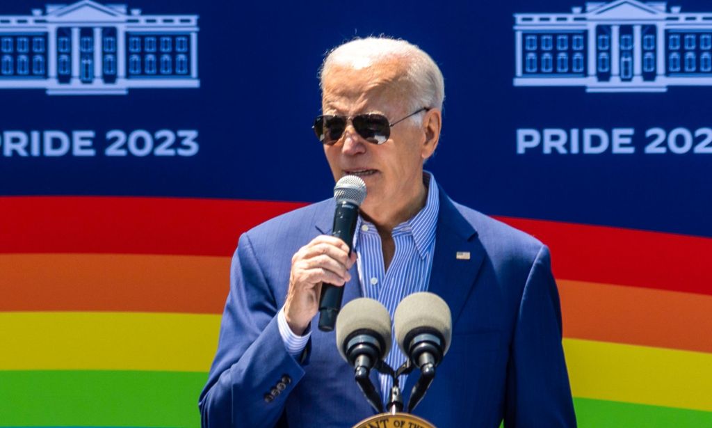 President Joe Biden speaking into a microphone, with a picture of a rainbow flag behind him.