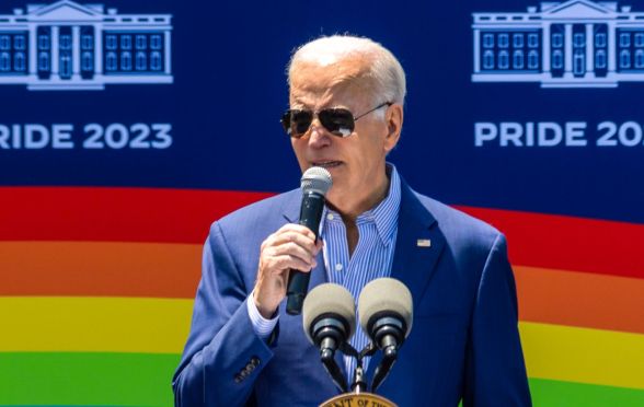 President Joe Biden speaking into a microphone, with a picture of a rainbow flag behind him.