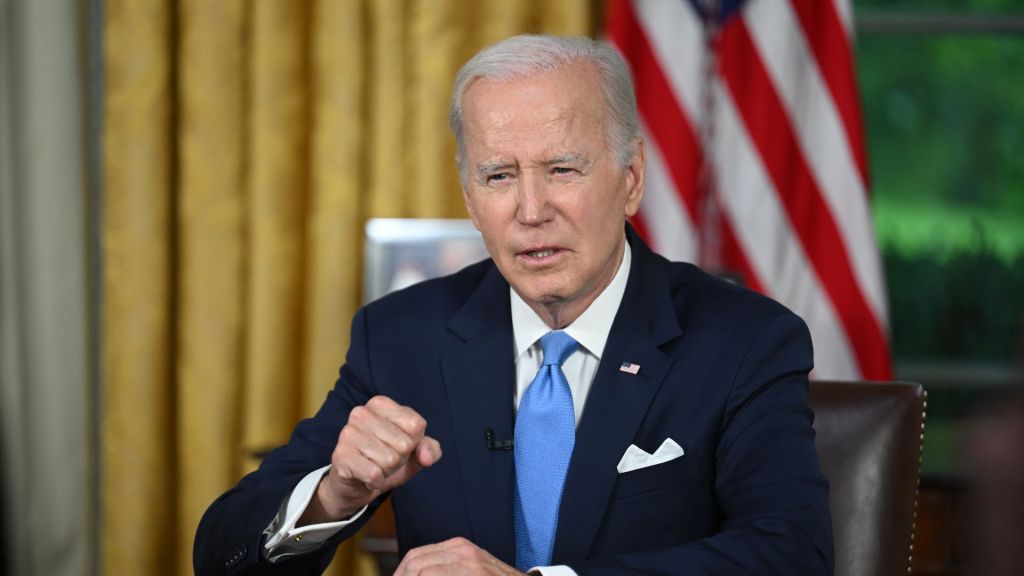 US President Joe Biden sits at a desk with an American flag behind him, delivering a talk from the White House