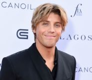 Lukas Gage in a black suit and shirt with parted blonde hair, smiling at the camera.