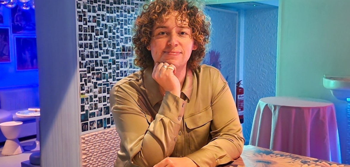 In this photo, Campaign Magazine's first transgender columnist Marty Davies is seen with curly brown hair and a khaki coloured shirt leaning on a table.
