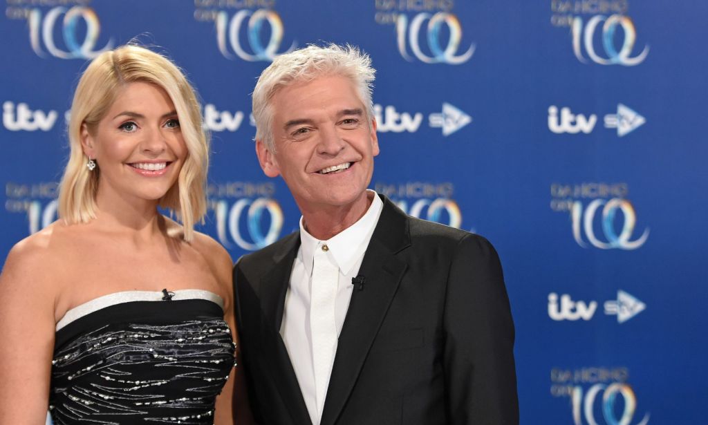 Holly Willoughby wears a black dress as she stands alongside Phillip Schofield as they stand in front of a blue background