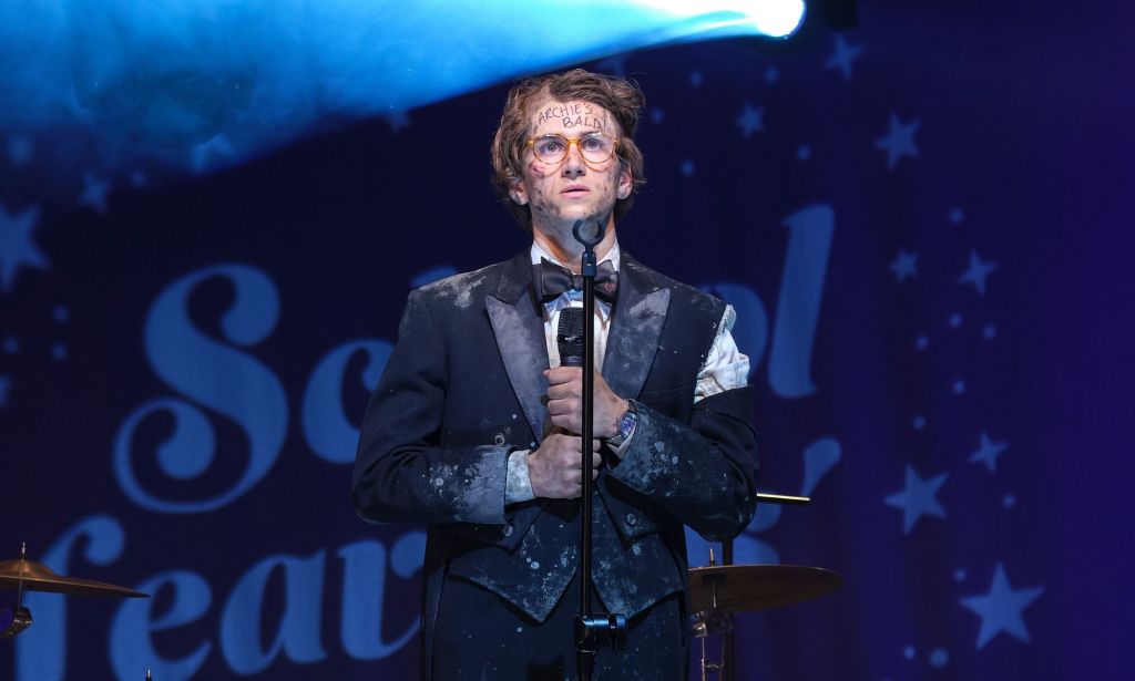 Sebastian Croft wears a filthy suit while standing on a stage and holding a microphone in a new still from new film How To Date Billy Walsh.