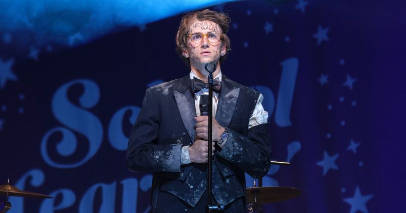 Sebastian Croft wears a filthy suit while standing on a stage and holding a microphone in a new still from new film How To Date Billy Walsh.