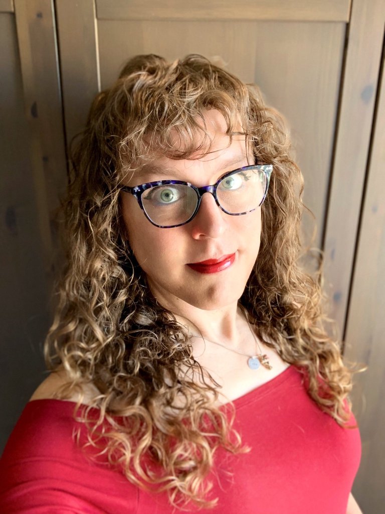 Tilly, a white woman with curly brown hair who is wearing glasses, red lipstick and a red dress