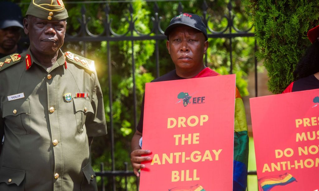 Protestors stand next to a uniformed officer, holding red signs that read "drop the anti-gay bill."