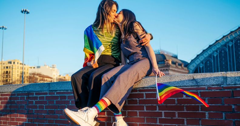 Photo of two women sitting on a wall kissing - one woman is holding a small Pride flag