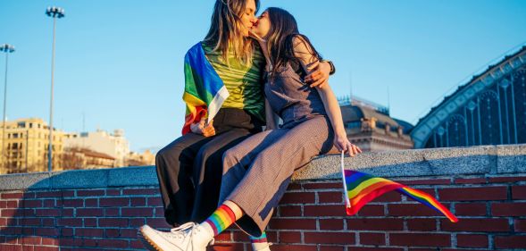 Photo of two women sitting on a wall kissing - one woman is holding a small Pride flag