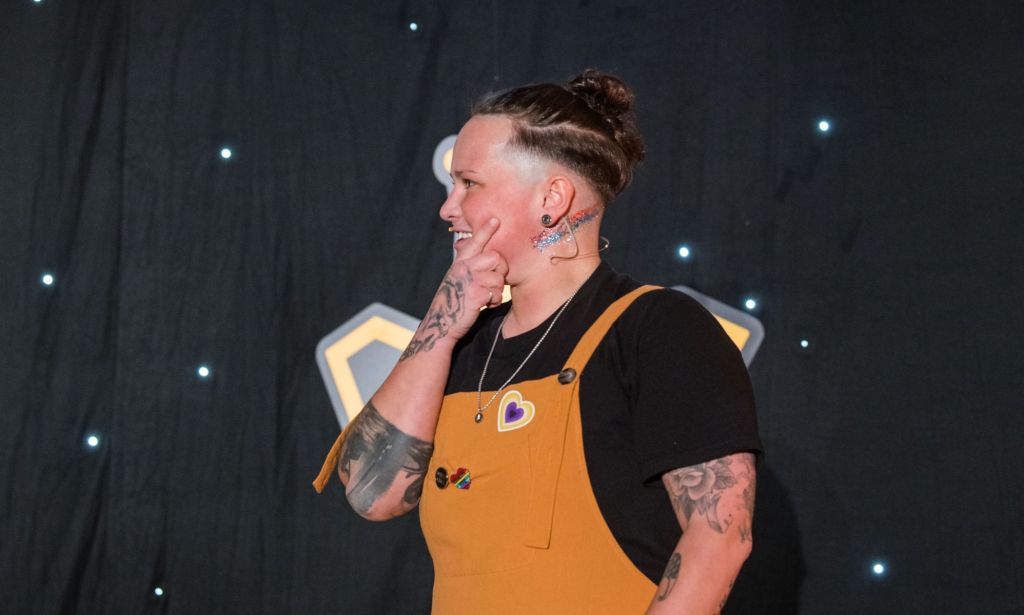 Non-binary speaker Alex Lynam wears a black top, yellow dungarees with a heart shaped pin in the colours of non-binary flag on their chest and a trans Pride flag glittered on their neck