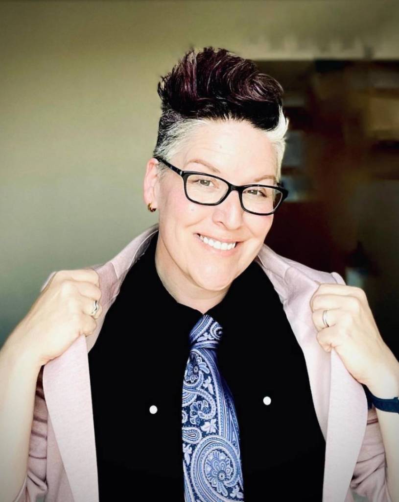 Author and advocate Amanda Jetté Knox wears a dark shirt, blue tie with a paisley pattern and light jacket which they are holding up by the labels as they tour around the UK to talk about trans rights and the power of allyship