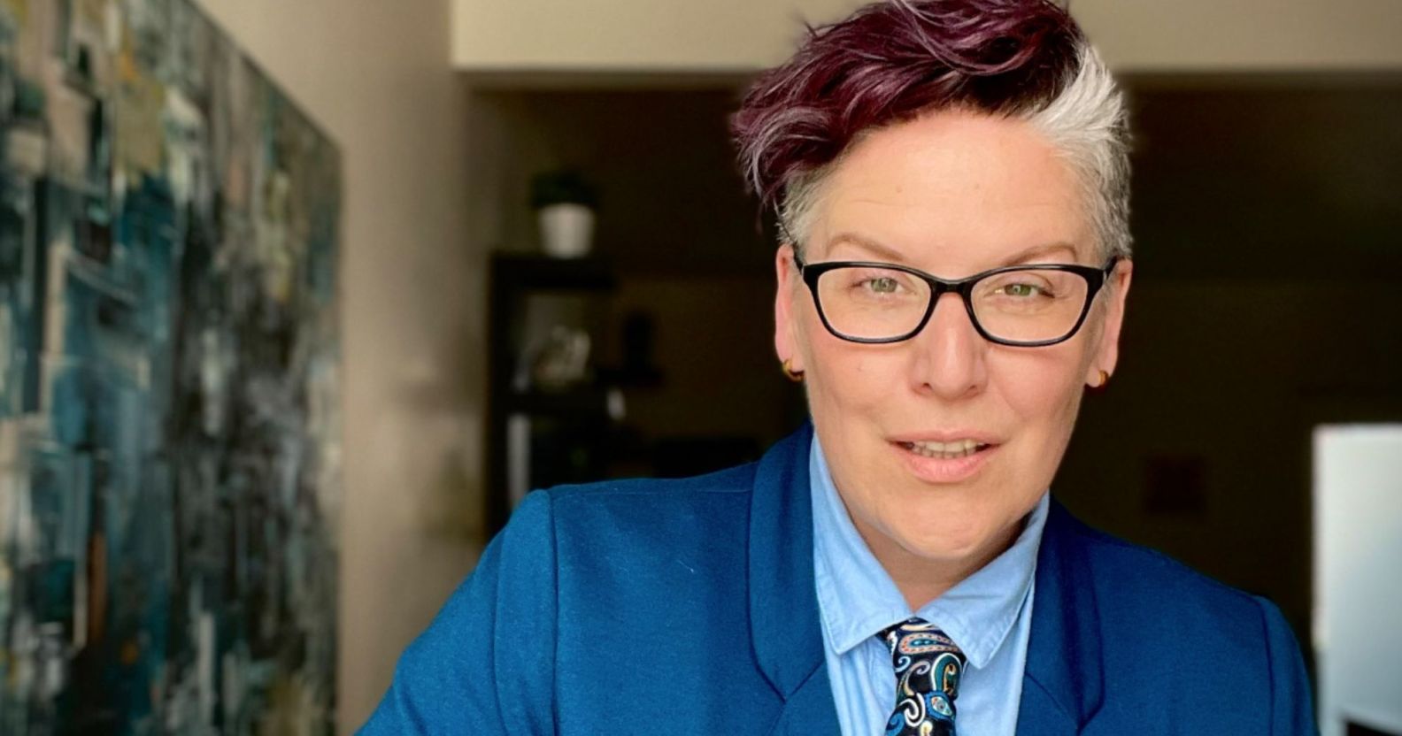 Author and advocate Amanda Jetté Knox wears a blue shirt, dark blue tie with a paisley pattern and medium toned blue jacket as they pose for the camera during their tour around the UK to talk about trans rights and the power of allyship