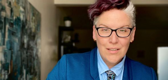 Author and advocate Rowan Jetté Knox wears a blue shirt, dark blue tie with a paisley pattern and medium toned blue jacket as they pose for the camera during their tour around the UK to talk about trans rights and the power of allyship