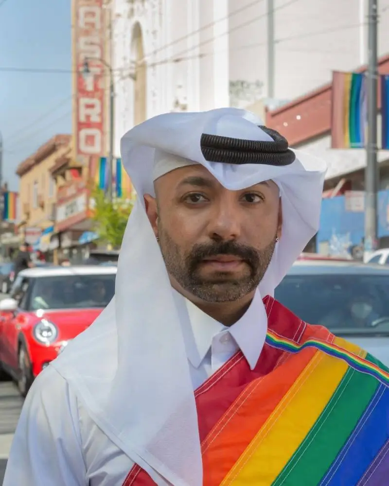Gay Qatari activist Dr Nasser Mohamed wears a white outfit and rainbow LGBTQ+ pride flag sash as he walks down a street