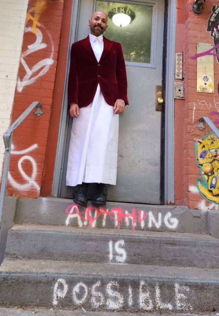 LGBTQ+ rights activist Dr Nasser Mohamed, who is from Qatar, wears a white outfit as he stands on the steps to a building with the words 'Anything is possible' written on the concrete