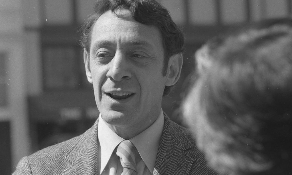 A black and white photo of Harvey Milk, who is wearing a suit and tie, as he speaks to people off camera