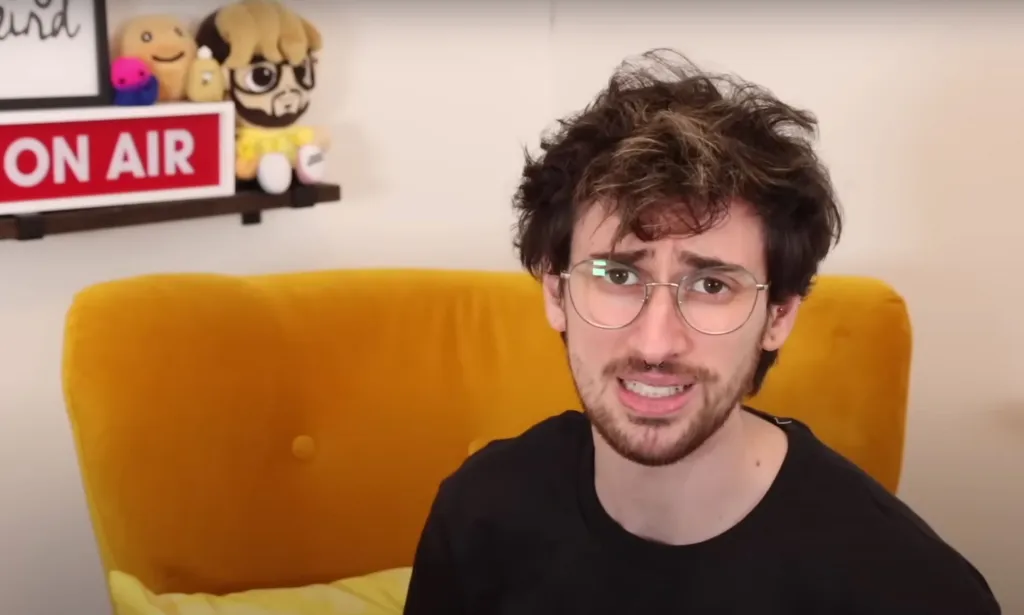 Jamie Raines, aka Jammidodger on YouTube, has dark brown hair and a matching beard. He is wearing a black shirt as he discusses topics relating to the trans community on his YouTube channel