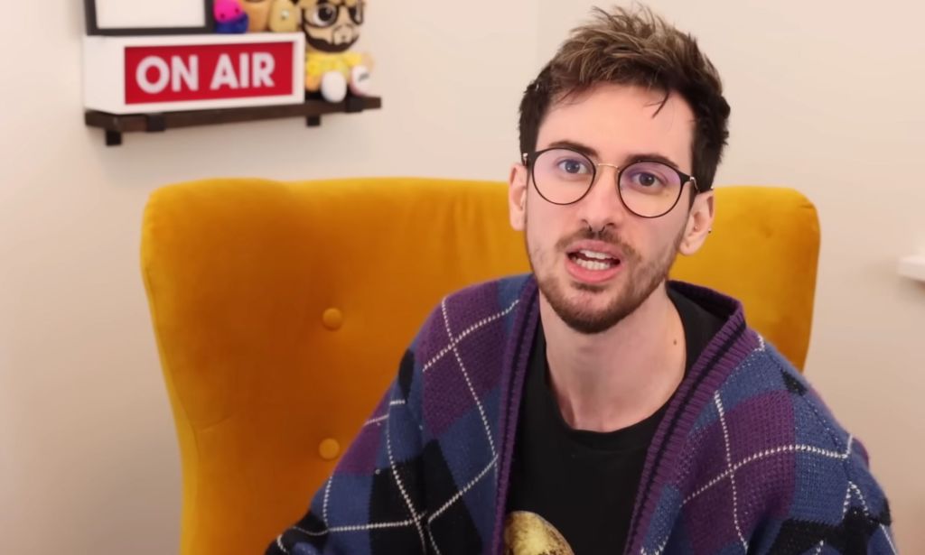 Jamie Raines, aka Jammidodger on YouTube, has dark brown hair and a matching beard. He is wearing a black shirt and purple and blue argyle print jumper as he discusses topics relating to the trans community on his YouTube channel