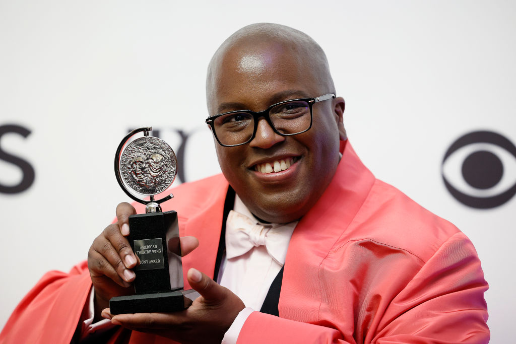 Michael, a Black man with a shaved head, wearing a long red coat, holding up a Tony award