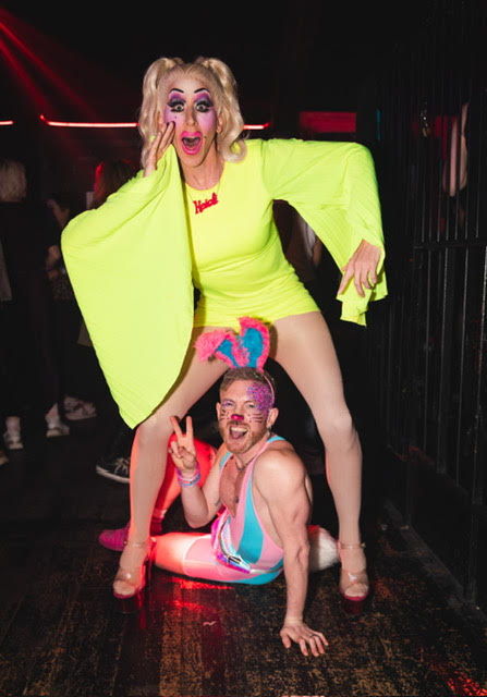A drag performer wears a yellow dress as a person wearing a pink and blue outfit poses on the ground between the performer's legs at a sober LGBTQ+ celebration