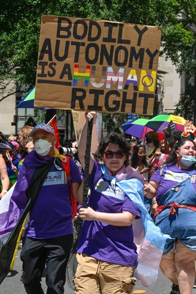 A person wearing a purple shirt holds up a sign reading 'bodily autonomy is a human right' with the word human designed in the colours of various LGBTQ+ and trans Pride flags during a protest