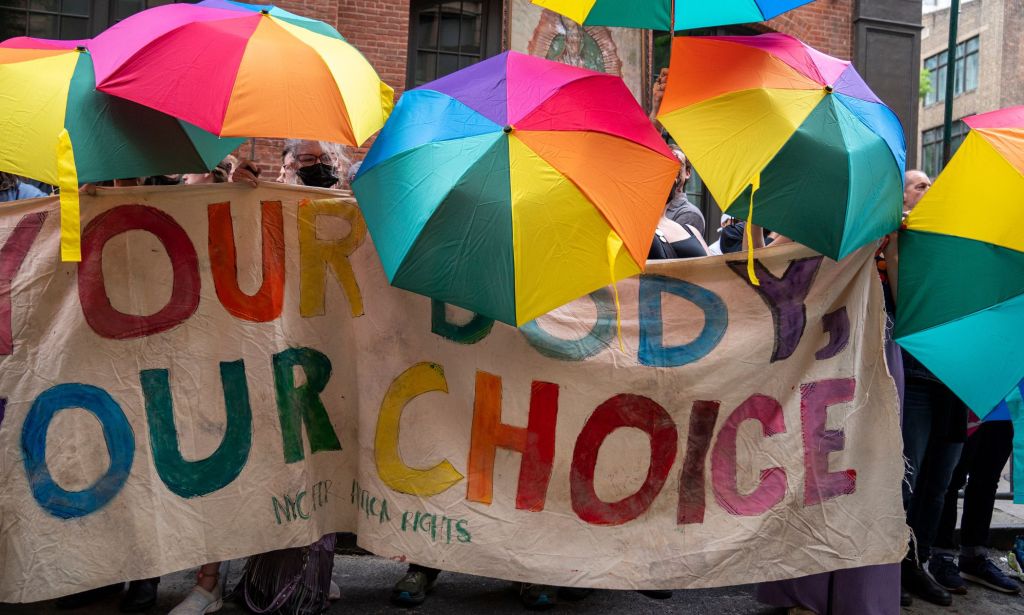 A crowd of people hold up rainbow coloured umbrellas with a sign reading 'your body your choice' during a protest composed of LGBTQ+ and trans people as well as allies against the Supreme Court overturning Roe v Wade 