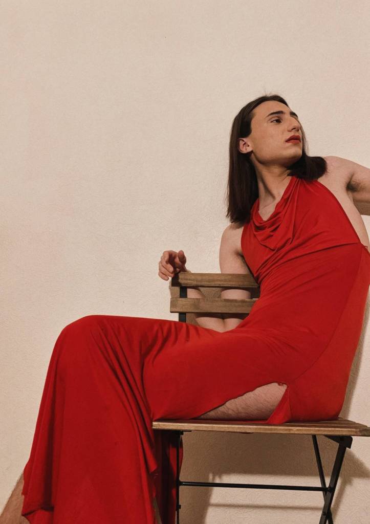 Mihelina, a Georgian trans woman living in Russia, has long dark brown hair and is wearing a red dress as she sits posed on a chair