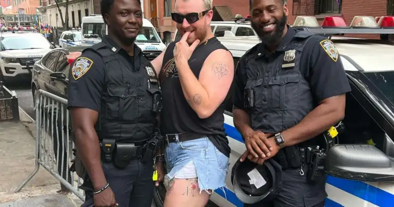 Sam Smith, a white non-binary person with blonde hair, posing in between two Black, male NYPD officers