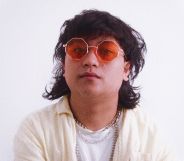 Non-binary, Malaysian singer Shaf (aka moreofthem) has shaggy dark hair and wears orange tinted glasses with a white shirt and off-white shirt on top with gold chains around their neck