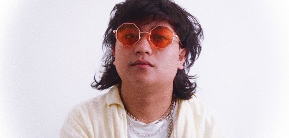 Non-binary, Malaysian singer Shaf (aka moreofthem) has shaggy dark hair and wears orange tinted glasses with a white shirt and off-white shirt on top with gold chains around their neck