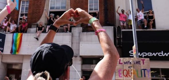 A person holds up their hands in the shape of a heart, to show their love, during an LGBTQ+ Pride event with a trans flag in the background