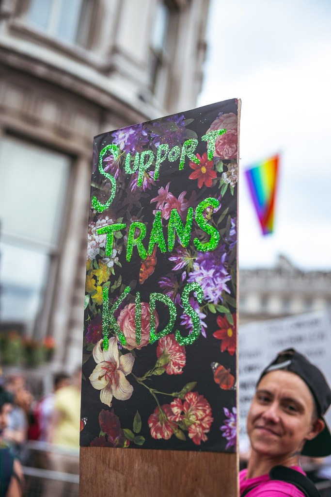A person holds up a sign reading 'support trans kids' during a protest with an LGBTQ+ flag seen in the background