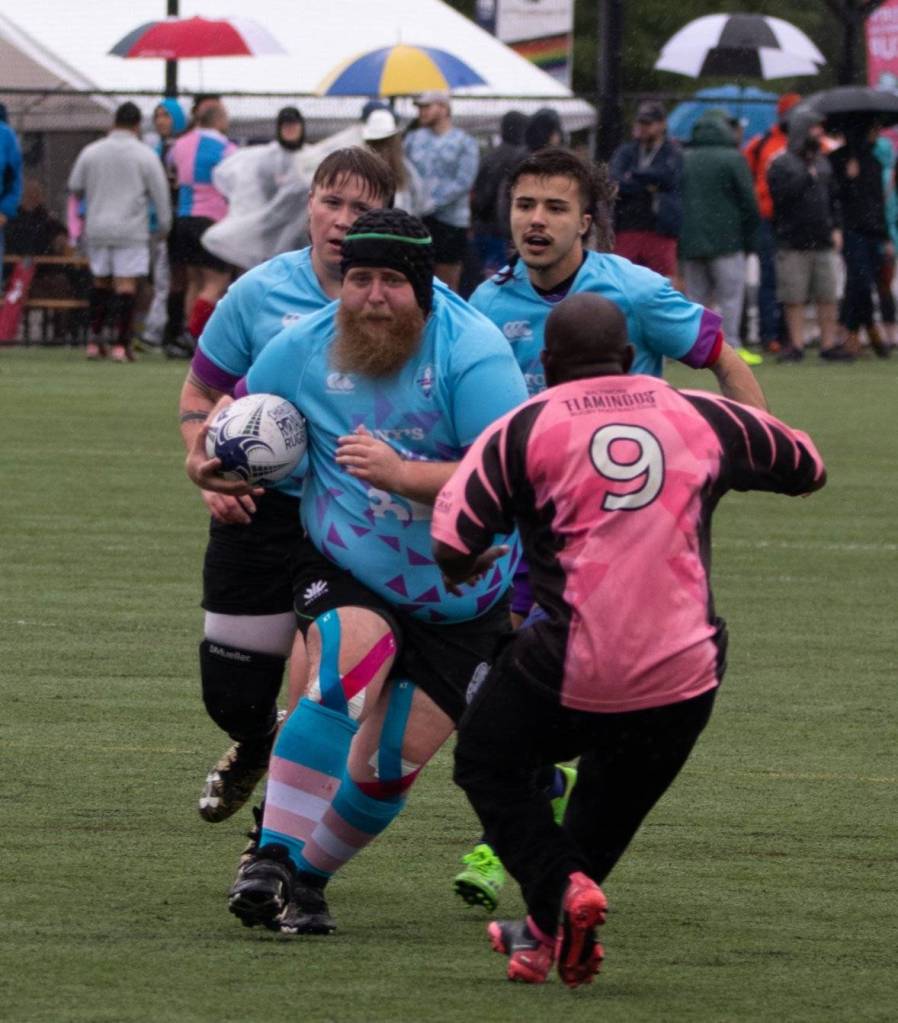 Trans player Atticus Martin wears a blue jersey as he runs with the ball during a rugby match as a player in pink runs to intercept him
