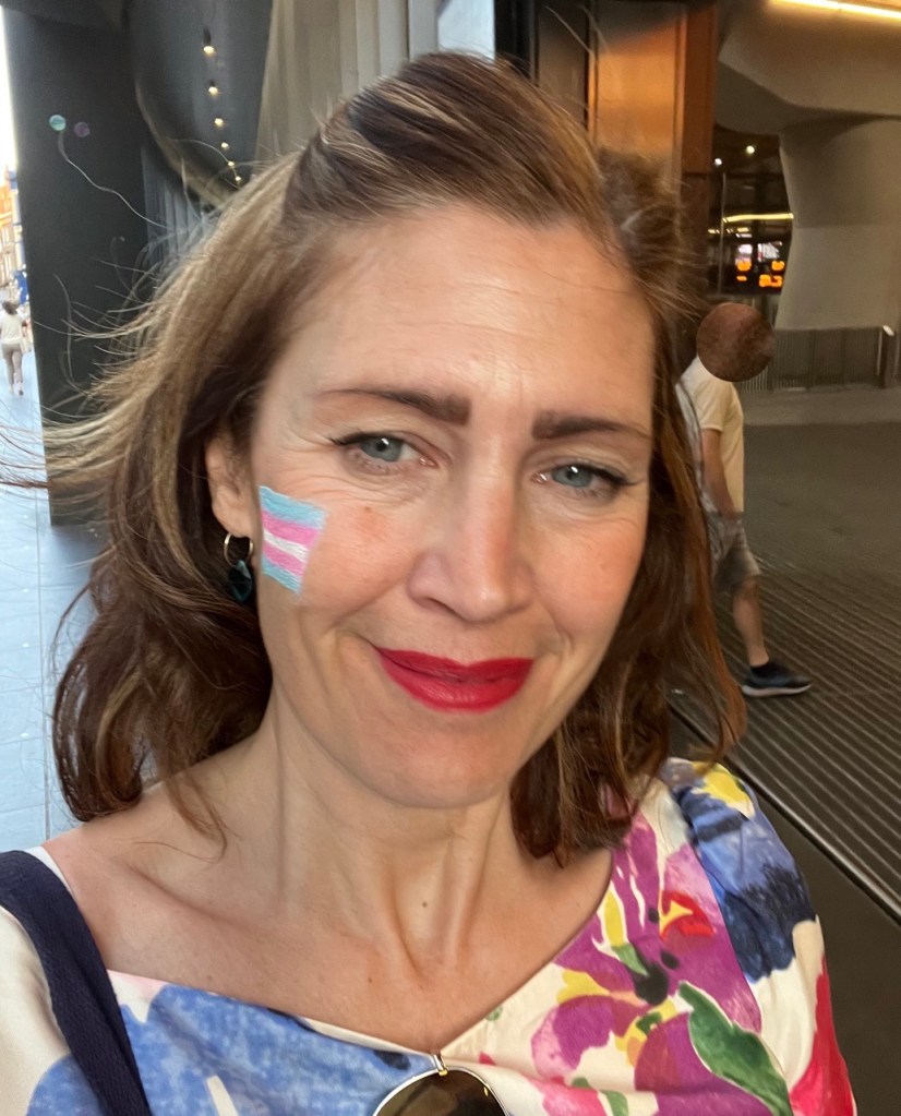 Aby Hawker, CEO and founder of TransMission PR, a communications consultancy which specialises in trans and non-binary inclusion and awareness, wearing a flower patterned dress with trans Pride face paint on her cheek