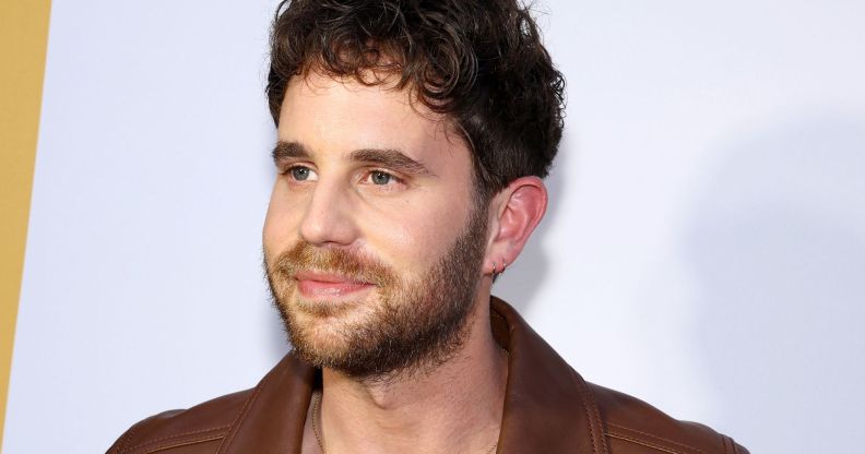 Ben Platt, wearing a brown jacket, smiles infront of a white background.